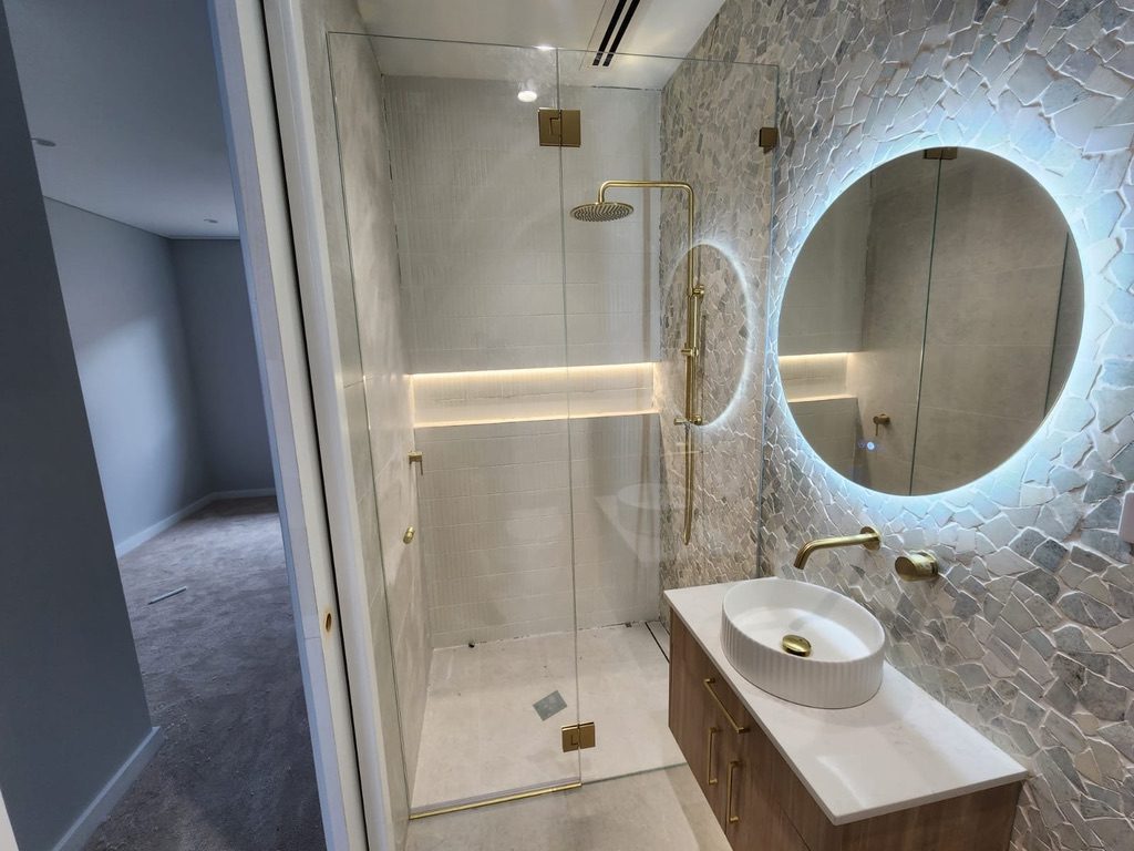 Frameless shower with brushed gold hardware and lit round stone-wall mirror