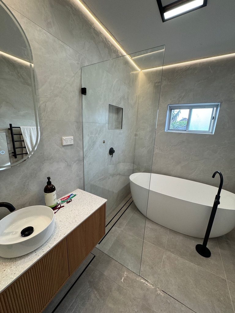 Frameless walk-in shower panel beside a freestanding bath with black tapware