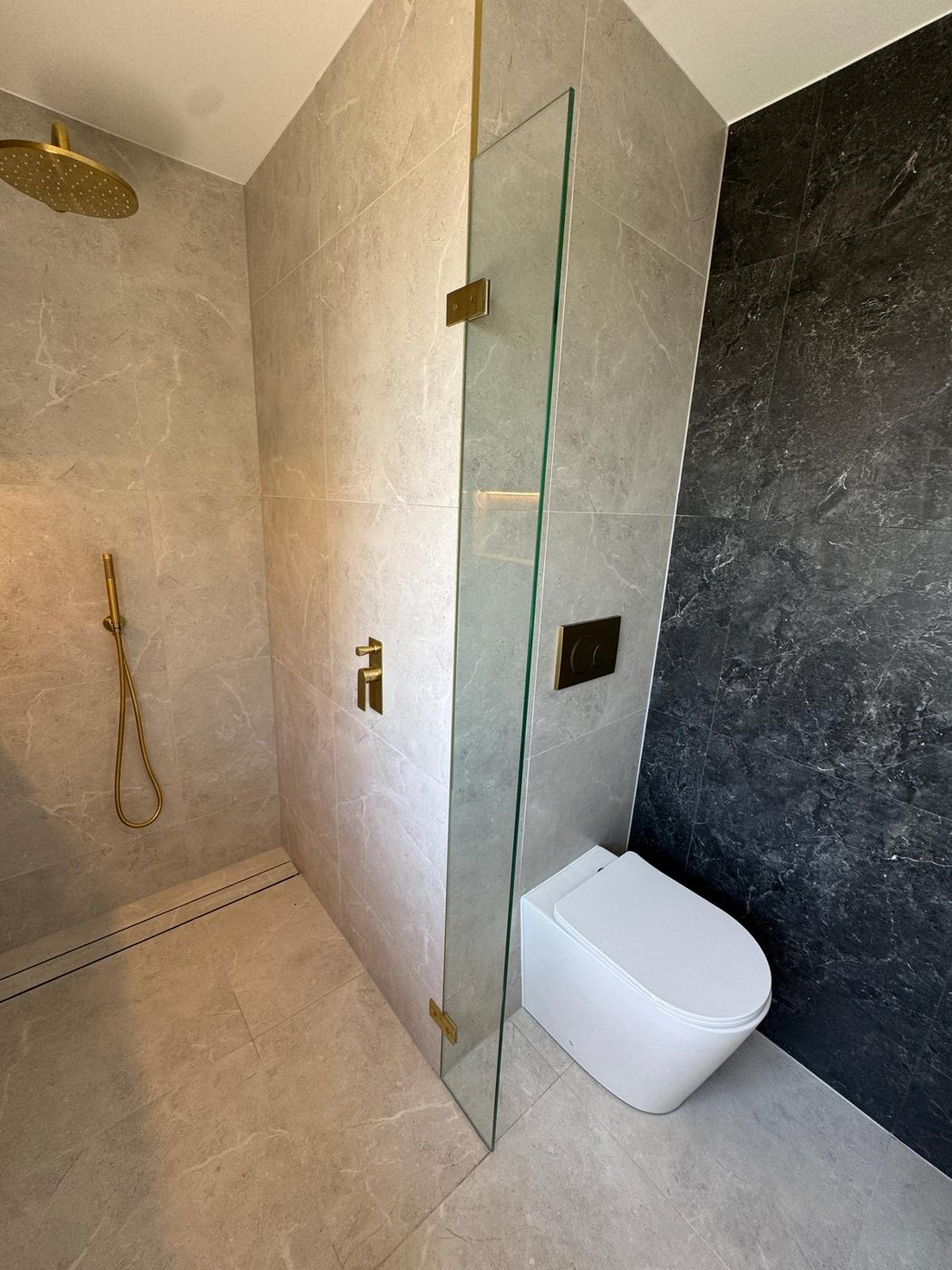 Frameless walk-in shower between grey and charcoal stone walls with brushed gold